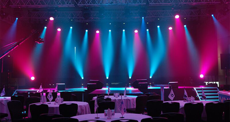 Die Bühne ist in einem leeren Raum mit violetten und blauen Lichtern beleuchtet. Im Vordergrund stehen runde Tische mit Stühlen und kleinen Fahnen. Darüber stützt ein Metallbalken die Beleuchtungskörper und schafft eine dramatische Atmosphäre.