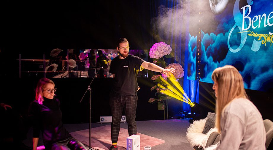 The person stands on stage and is in contact with the audience. Facing them are two seated individuals, surrounded by mood lighting and a large screen with a sign that reads „Benefit“. Decorative elements such as flowers and fog accentuate the scene.