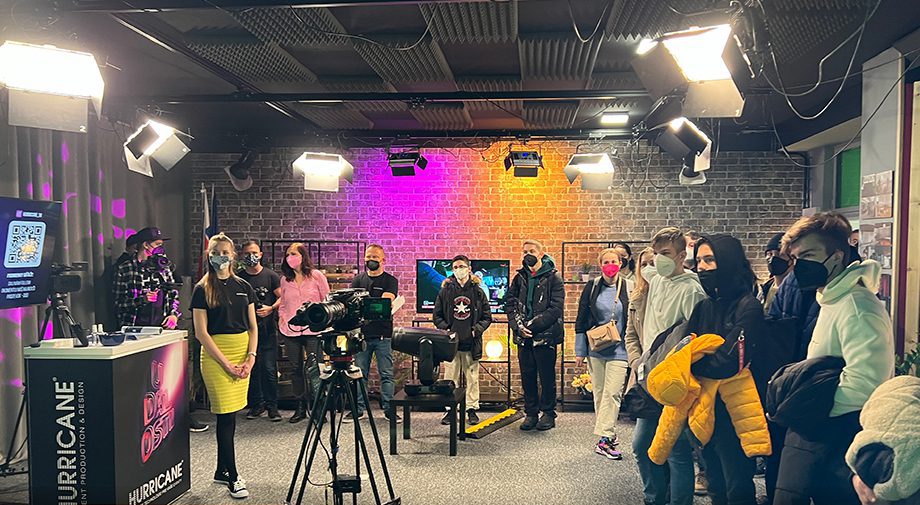 Eine Gruppe von Personen, von denen einige Masken tragen, steht in einem hell erleuchteten Studio mit Kameras und Beleuchtungsausrüstung. Eine Backsteinmauer mit farbigen Lichtern ziert den Hintergrund. Einige Personen halten Mäntel in ihren Armen.