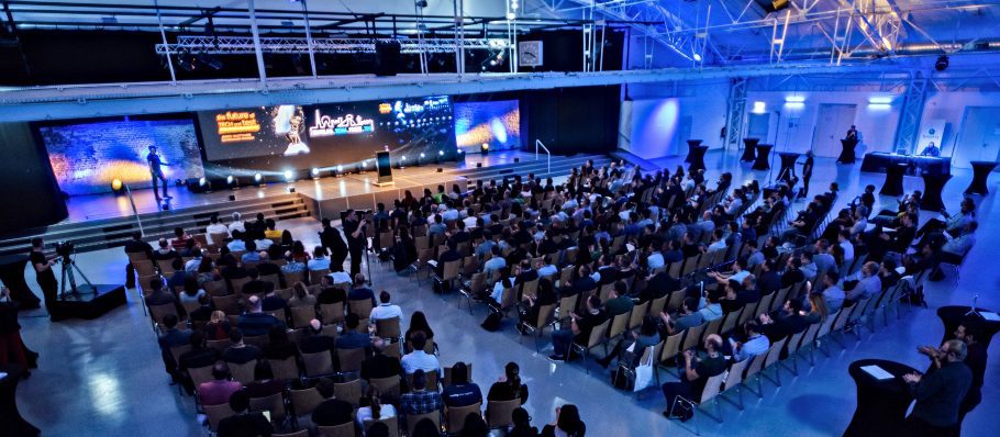 Großes Publikum, das in Reihen vor der Bühne der Konferenz sitzt. Auf der Bühne befinden sich ein Redner und mehrere Bildschirme, auf denen Grafiken angezeigt werden. Der Raum ist geräumig mit hohen Decken und heller Beleuchtung.