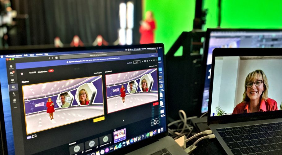 Arbeitsbereich mit zwei Laptops. Einer zeigt eine Videokonferenzanwendung mit einem virtuellen Podium und zwei identische Bildschirme mit einer Moderatorin. Der andere Laptop zeigt eine lächelnde Frau in einem kleineren Videoanruffenster. Ein heller grüner Bildschirm im Hintergrund.