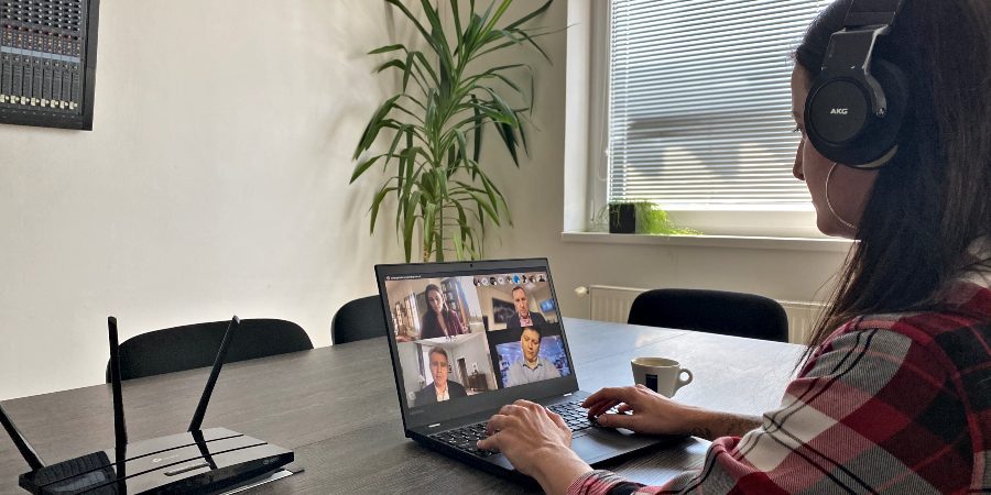 Eine Person mit Kopfhörern sitzt an einem Tisch und nimmt an einem Videogespräch über einen Laptop teil. Auf dem Bildschirm sind mehrere Personen zu sehen. Eine Tasse Kaffee, ein Router und eine große Pflanze stehen in der Nähe in einem hellen Raum.