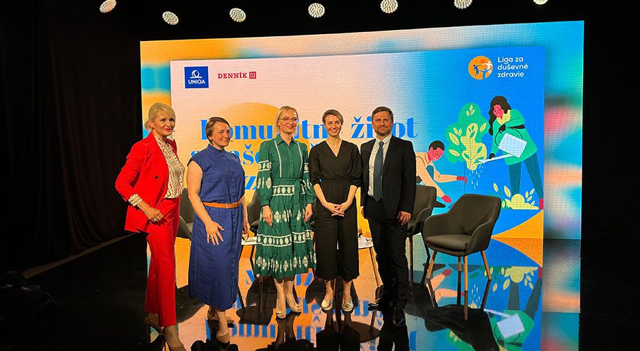 Eine Gruppe von fünf Personen steht auf einer Bühne vor einem bunten Hintergrund mit Illustrationen und Text. Die Personen sind formell gekleidet, zwei Männer in Anzügen und drei Frauen in Kleidern. Neben ihnen stehen zwei leere Stühle.