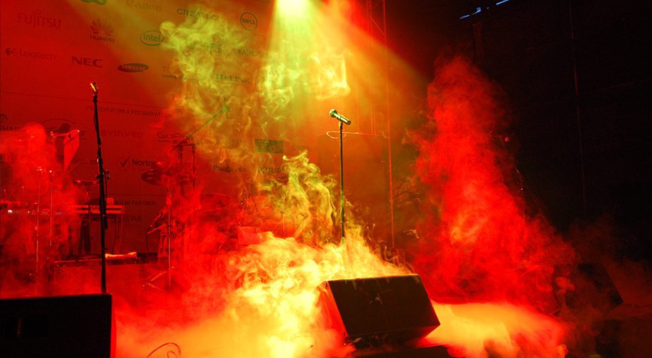 Eine Live-Konzertbühne mit wirbelndem rotem und orangefarbenem Rauch, beleuchtet von hellen Lichtern. Der Mikrofonständer steht in der Mitte, und im Hintergrund sind verschiedene Logos zu sehen, die auf eine energiegeladene Performance hinweisen.