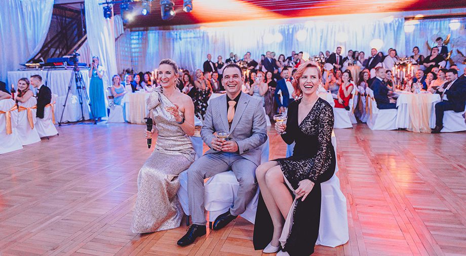 Drei festlich gekleidete Personen sitzen zusammen. Im Hintergrund sitzt ein großes Publikum an dekorierten Tischen und genießt die lebhafte Veranstaltung in einem hell erleuchteten Raum mit Wänden.