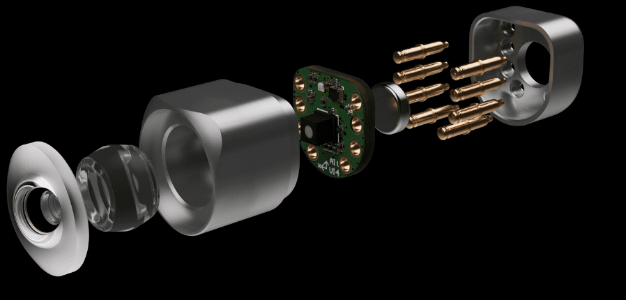 Exploded view of the mechanical device showing the individual components including the metal housing, cylindrical parts, circuit board and small brass rods, all against a black background.