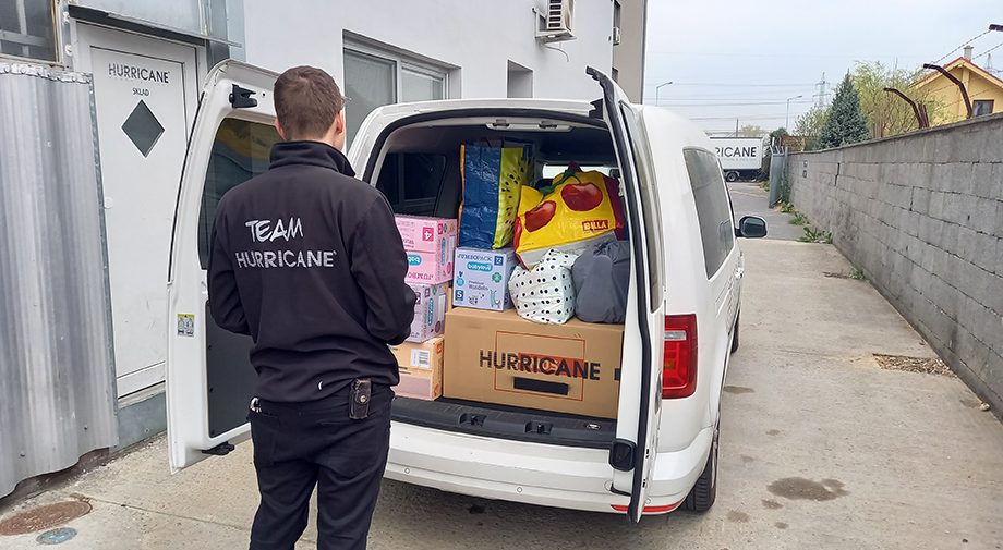 Eine Person, die eine „Team Hurricane“-Jacke trägt, steht vor einem offenen weißen Lieferwagen, der mit verschiedenen Kisten und Paketen beladen ist. Der Lieferwagen ist auf einer Betonfläche neben einem Gebäude mit der Aufschrift „Hurricane“ geparkt.