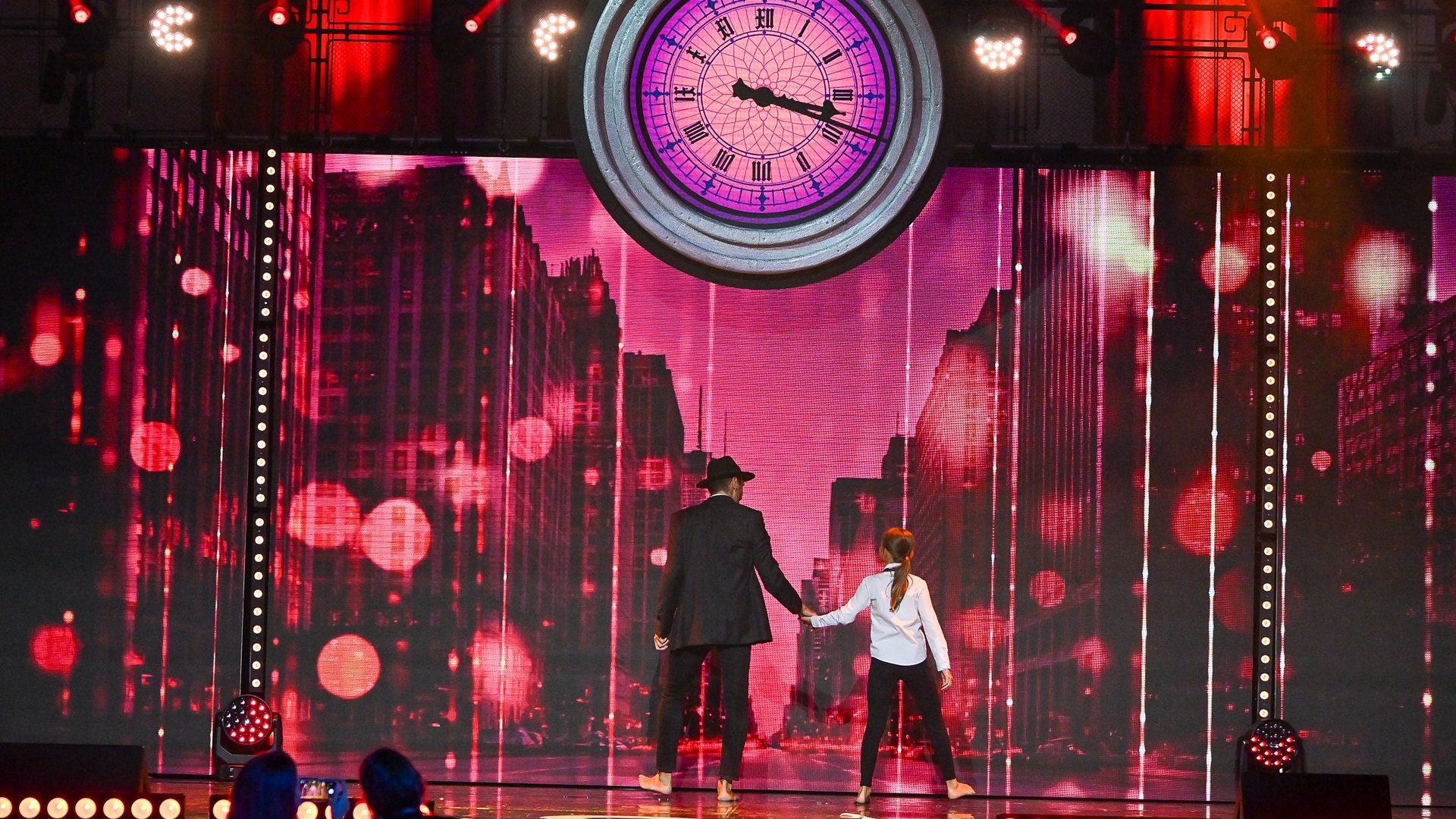 Ein Paar auf der Bühne vor einer Stadtkulisse mit einer Uhr.