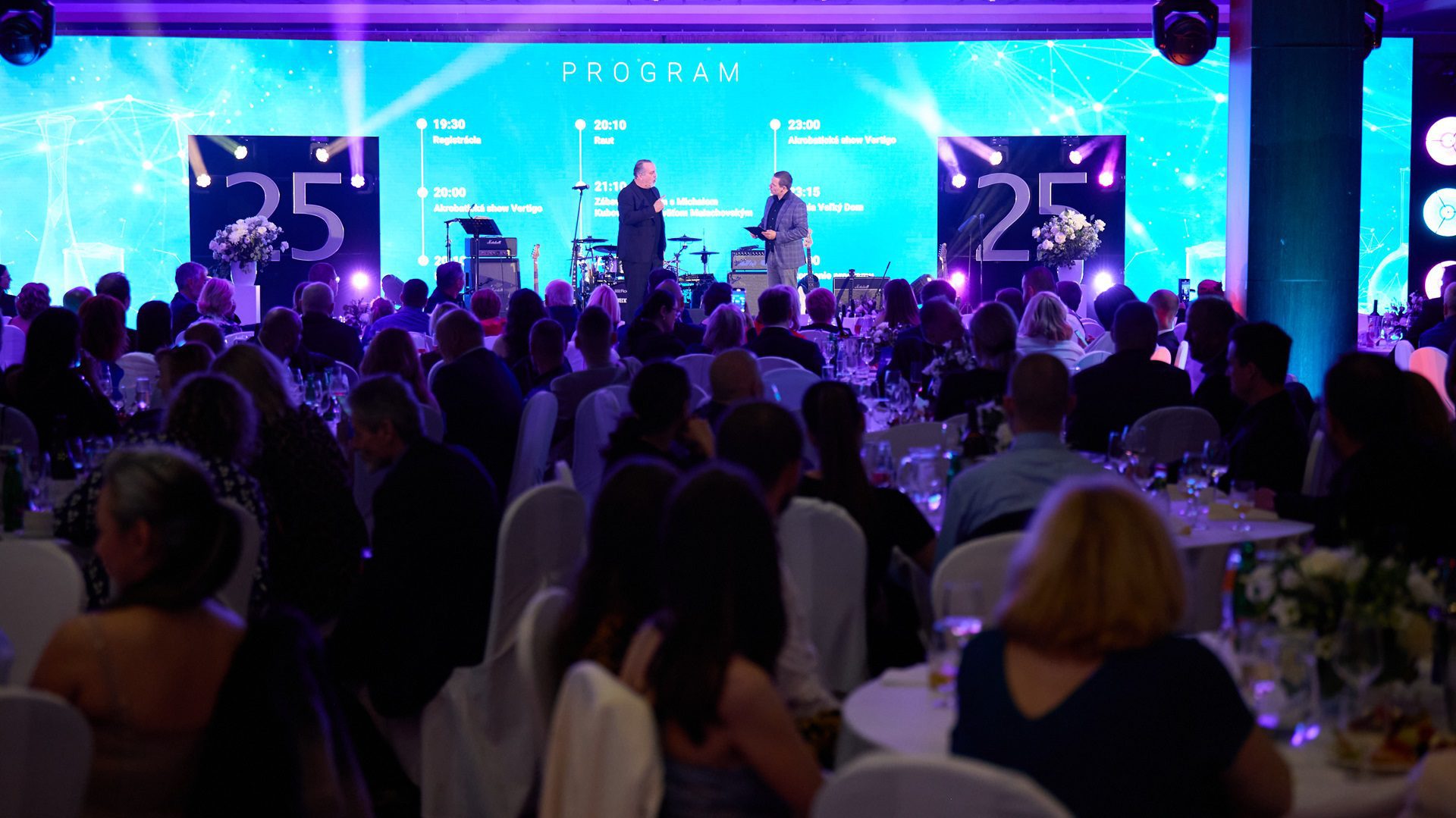 Ľudia na podujatí s rečníkmi na pódiu.