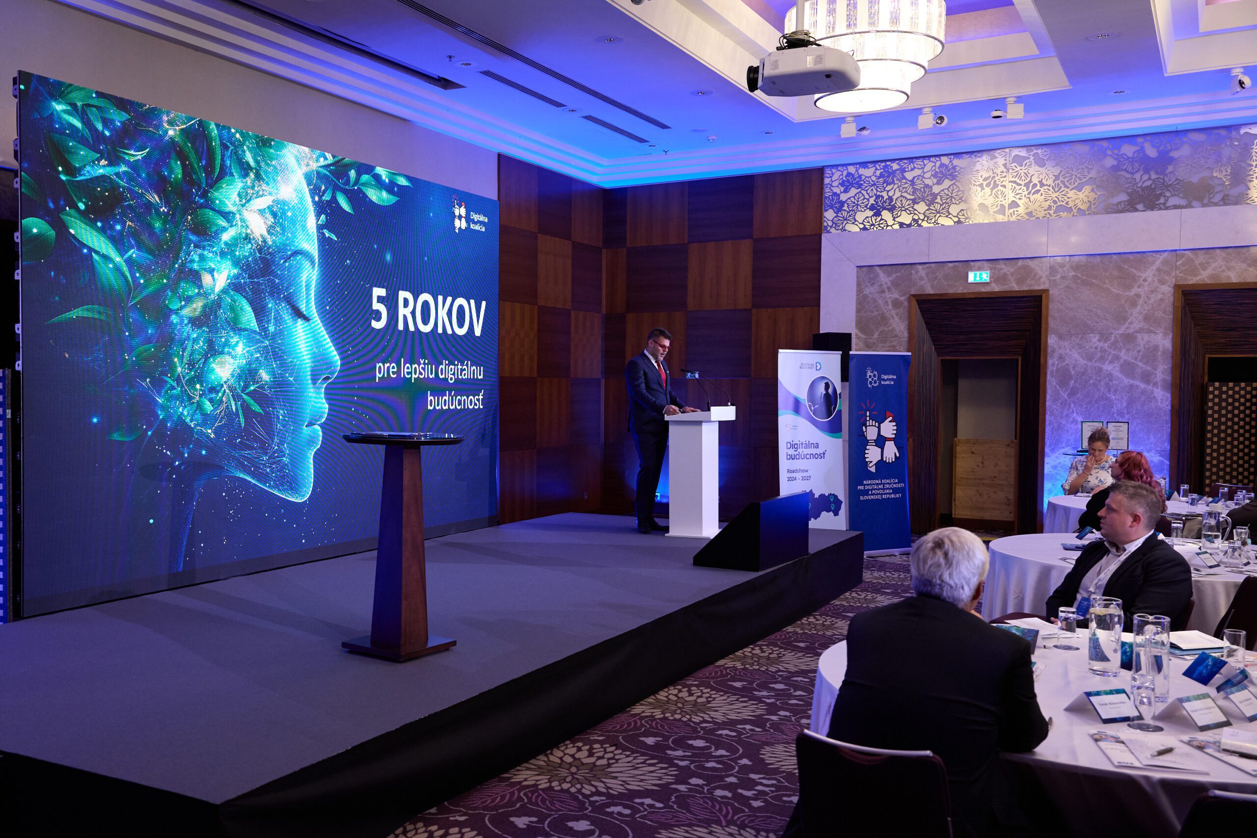 Der Redner steht auf der Bühne und präsentiert vor einem großen LED-Bildschirm mit dem Thema der digitalen Zukunft.