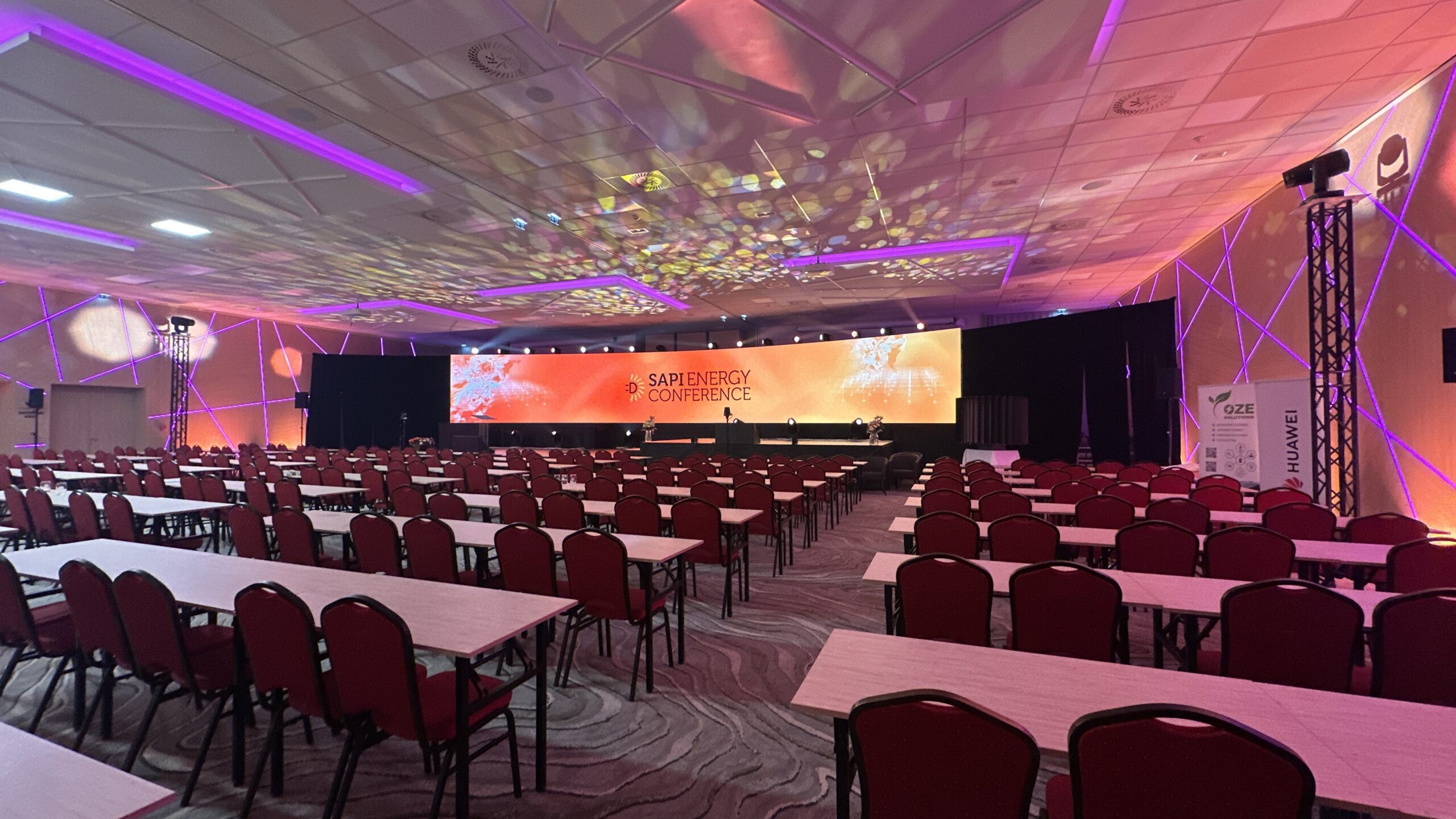 Konferenzraum mit Tischen und Stühlen, großer LED-Leinwand mit SAPI Energy Conference Logo und Lichtdesign an den Wänden