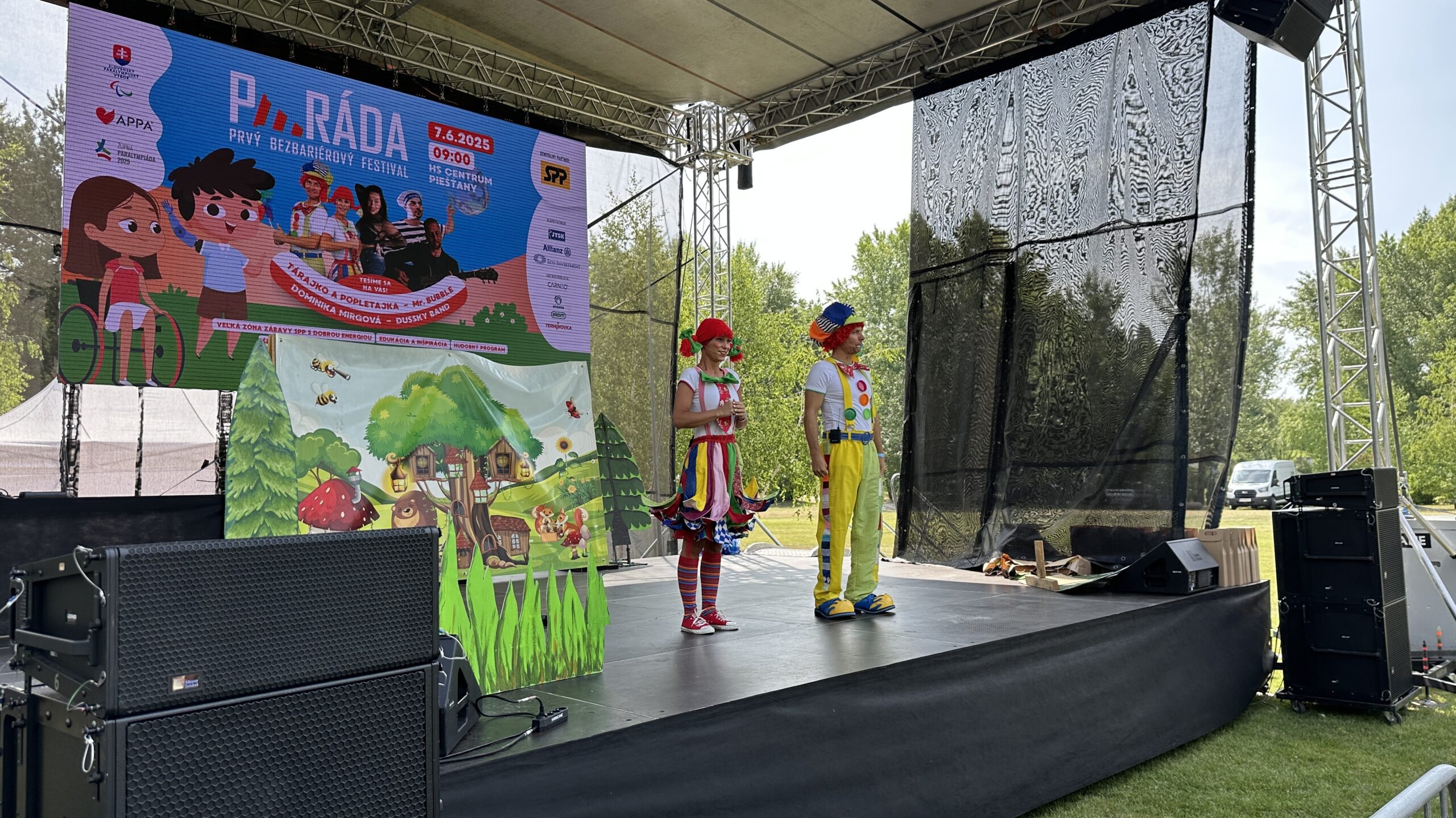 Dvaja klauni v pestrofarebných kostýmoch stoja na pódiu počas festivalu PÁRÁDA v Piešťanoch, v pozadí veľká obrazovka s programom podujatia a logami partnerov.