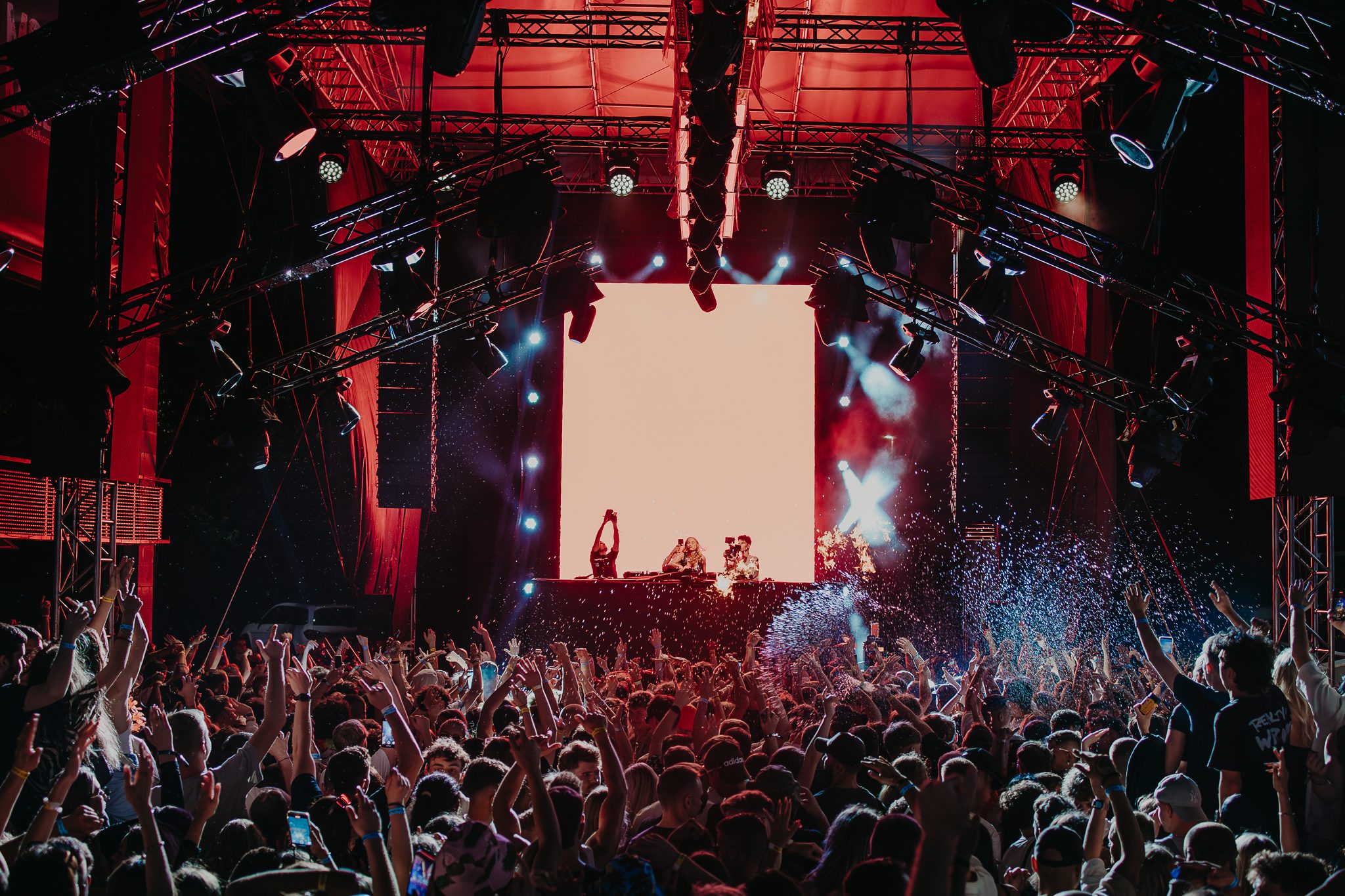 Große Menge von Fans mit den Händen in der Luft vor der Bühne mit DJ, roter Beleuchtung, Lichtshow und visuellen Effekten auf dem Nachtfestival