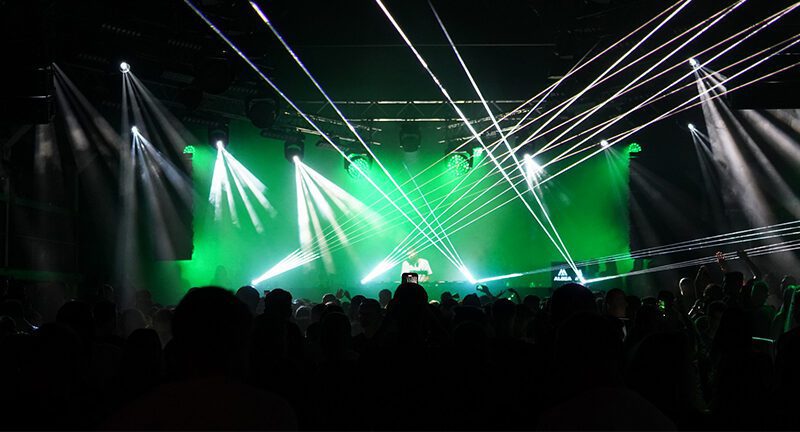 Scéna preplneného nočného klubu s farebnými laserovými svetlami žiariacimi cez miestnosť. Ľudia môžu vidieť, ako si užívajú atmosféru, s DJ stánkom osvetleným v pozadí uprostred zelených a bielych svetiel.