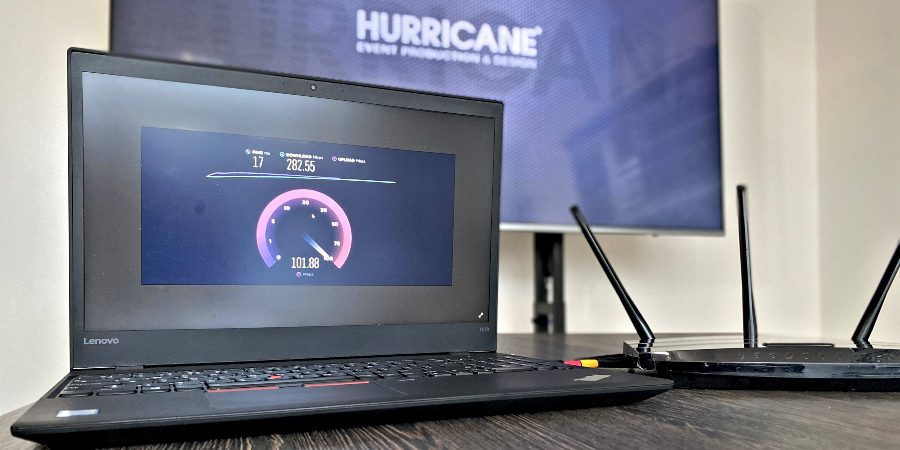 Prenosný počítač zobrazujúci výsledok testu rýchlosti internetu leží na stole vedľa smerovača Wi-Fi. V pozadí sa na televíznej obrazovke zobrazuje slovo „HURRICANE“ veľkým textom.