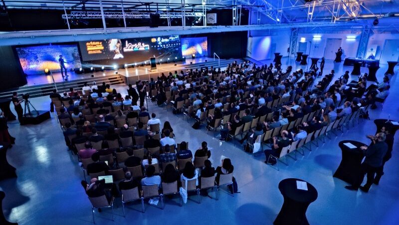 A large conference room full of people sitting on chairs across from a podium with a speaker and multiple screens with presentations. The room is well lit with blue ambient lighting. There are round black tables scattered around the back.