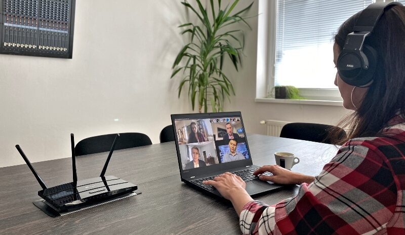 A person wearing headphones participates in a videoconference on a laptop computer at a table. A router with multiple antennas is nearby. In the background is a large plant and a window with blinds. Next to the laptop is a small mug.