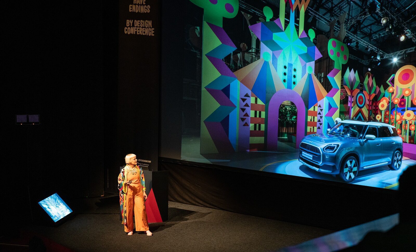 Žena na pódiu na konferencii By Design pred veľkým projekčným plátnom s farebnou geometrickou scénografiou a modrým autom