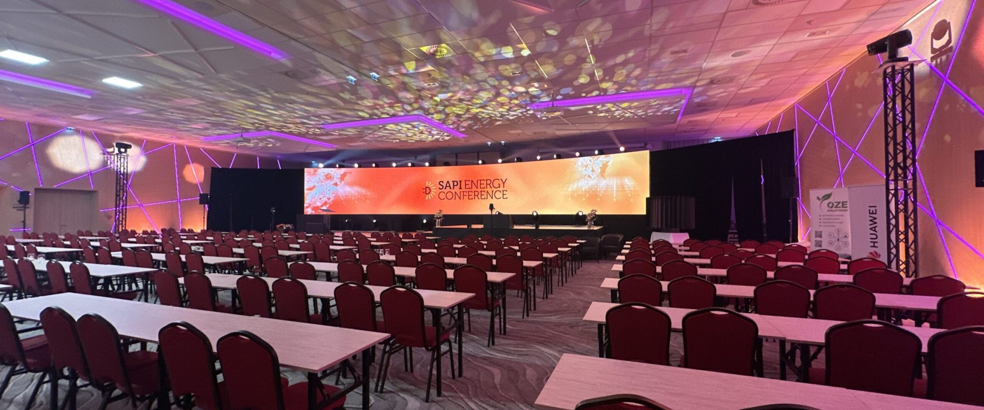 Konferenzraum mit Tischen und Stühlen, großer LED-Leinwand mit SAPI Energy Conference Logo und Lichtdesign an den Wänden