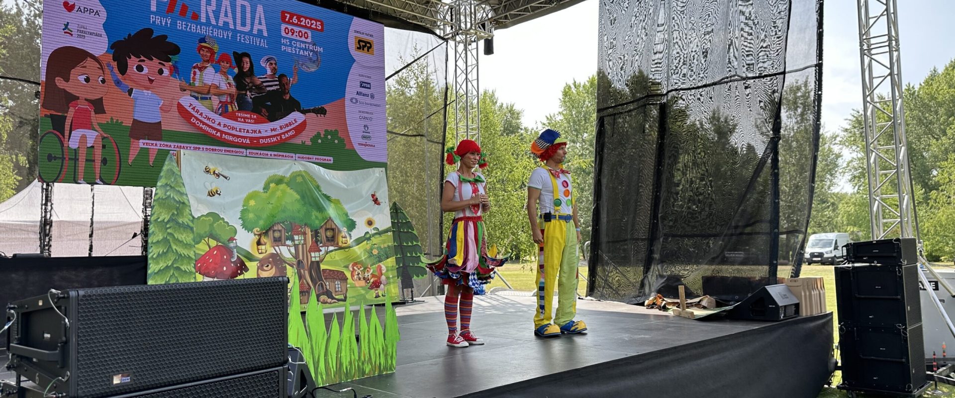 Dvaja klauni v pestrofarebných kostýmoch stoja na pódiu počas festivalu PÁRÁDA v Piešťanoch, v pozadí veľká obrazovka s programom podujatia a logami partnerov.