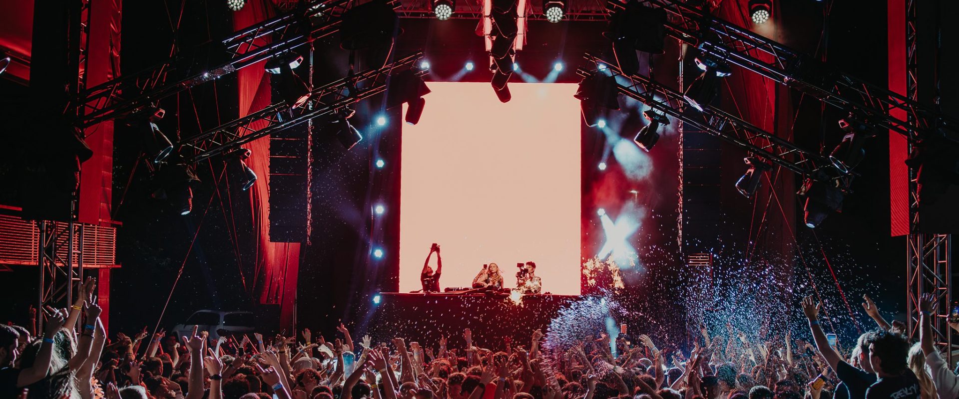 Große Menge von Fans mit den Händen in der Luft vor der Bühne mit DJ, roter Beleuchtung, Lichtshow und visuellen Effekten auf dem Nachtfestival