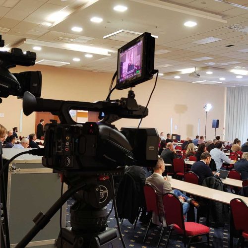 Konferenz in einem großen Saal mit einer Kamera im Hintergrund.