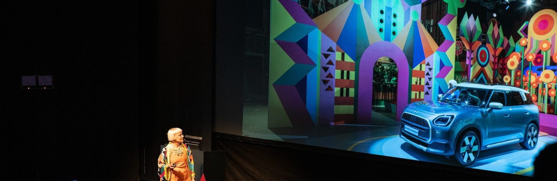 Eine Frau auf der Bühne der By Design-Konferenz vor einer großen Projektionsfläche mit einer farbenfrohen geometrischen Szenografie und einem blauen Auto