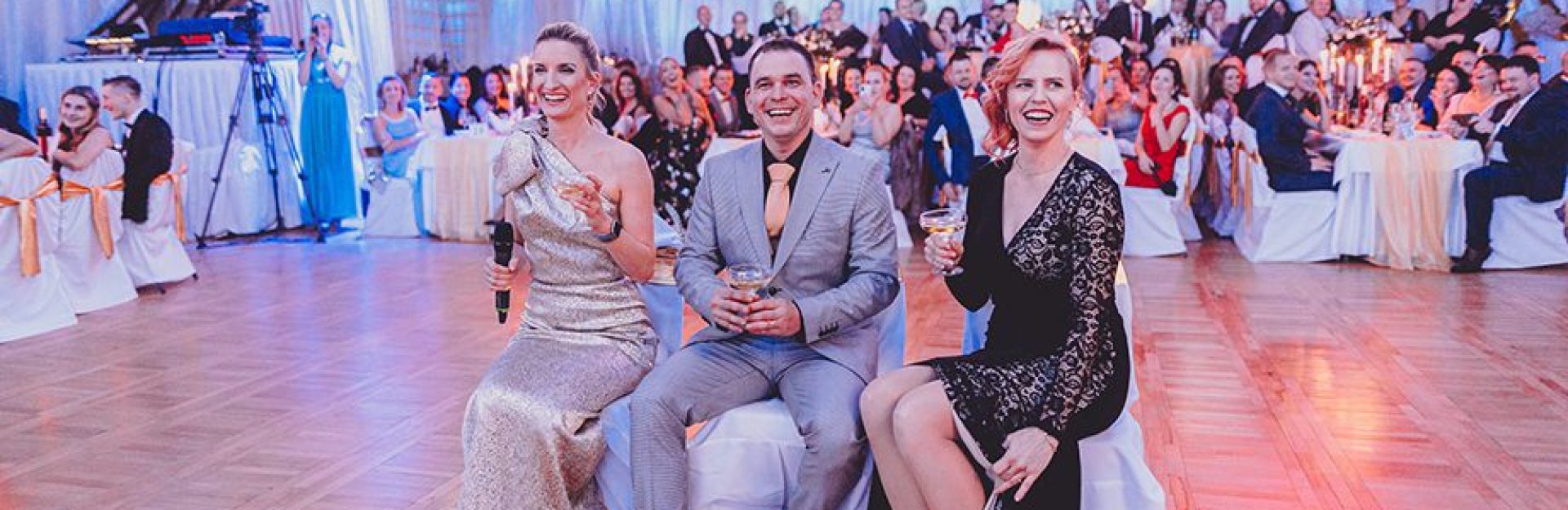 Drei festlich gekleidete Personen sitzen zusammen. Im Hintergrund sitzt ein großes Publikum an dekorierten Tischen und genießt die lebhafte Veranstaltung in einem hell erleuchteten Raum mit Wänden.
