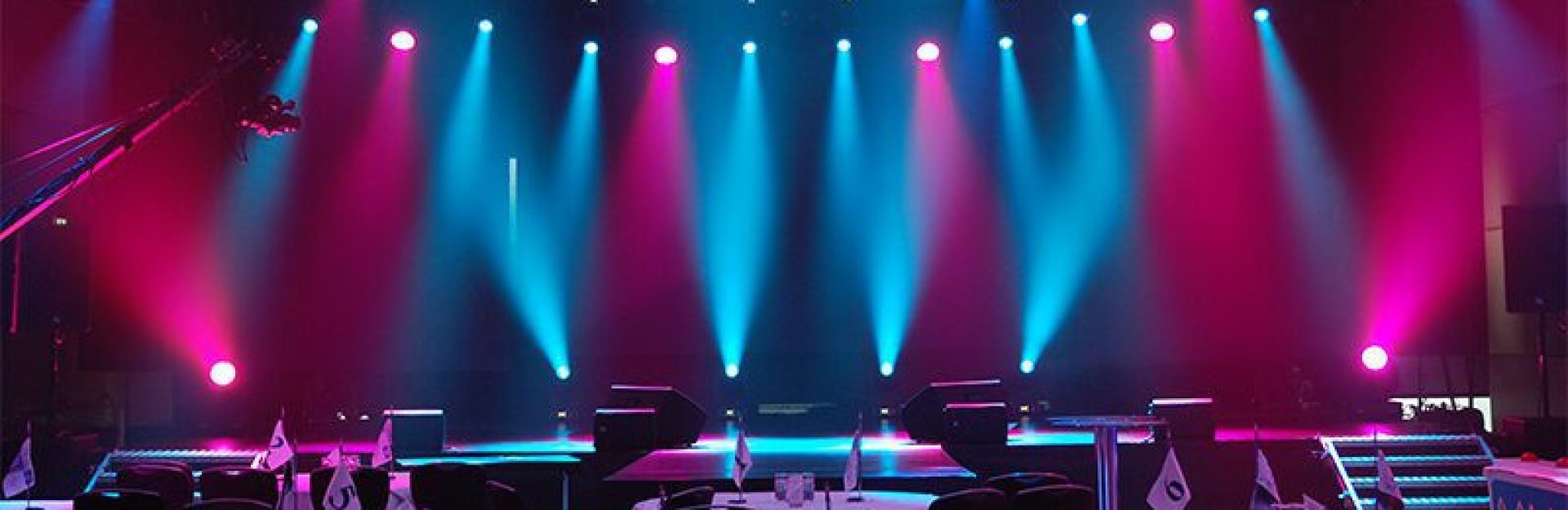 Die Bühne ist in einem leeren Raum mit violetten und blauen Lichtern beleuchtet. Im Vordergrund stehen runde Tische mit Stühlen und kleinen Fahnen. Darüber stützt ein Metallbalken die Beleuchtungskörper und schafft eine dramatische Atmosphäre.