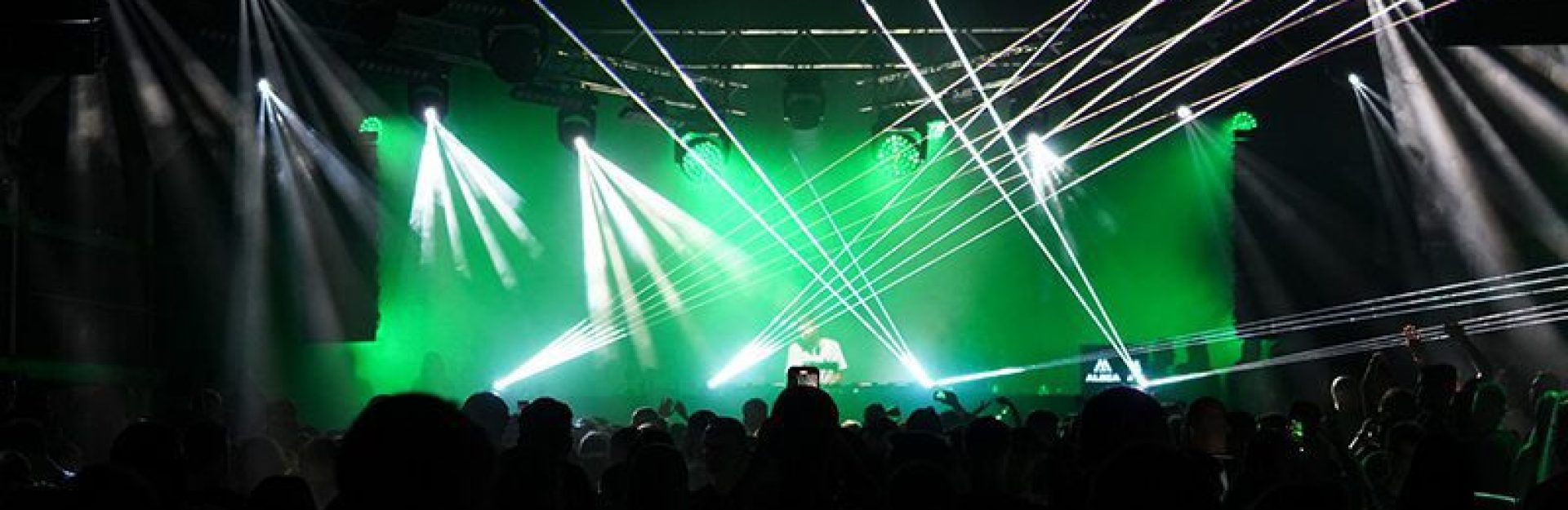 The scene of a crowded nightclub with colored laser lights shining across the room. People can be seen enjoying the atmosphere, with the DJ booth backlit amidst the green and white lights.