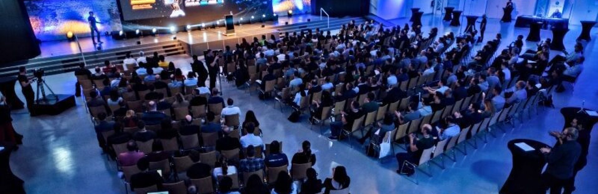 Ein großer Konferenzraum voller Menschen, die auf Stühlen gegenüber einem Podium mit einem Redner und mehreren Bildschirmen mit Präsentationen sitzen. Der Raum ist gut beleuchtet mit blauer Umgebungsbeleuchtung. Im hinteren Teil sind runde schwarze Tische aufgestellt.