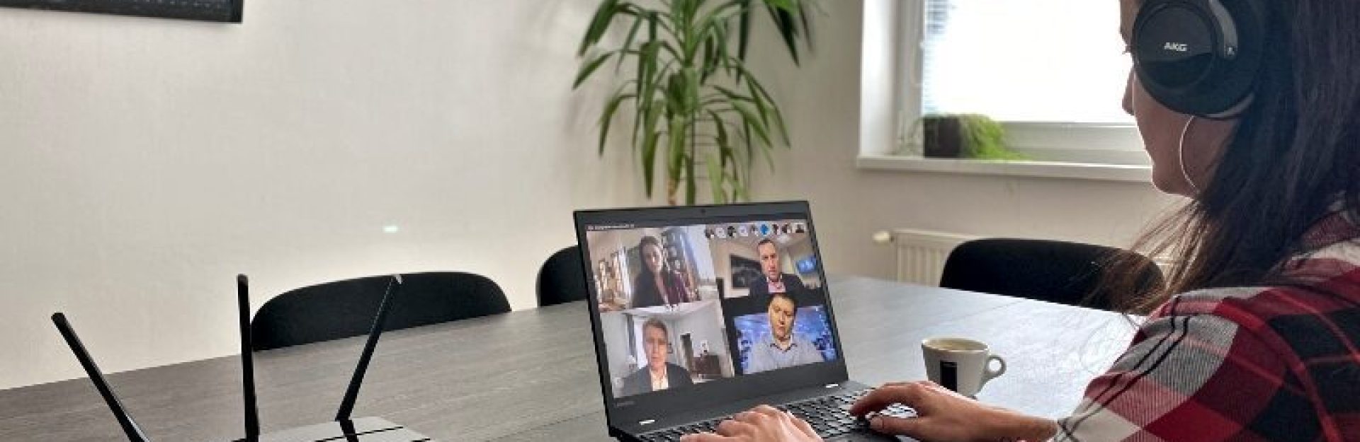 Eine Person, die Kopfhörer trägt, nimmt an einer Videokonferenz auf einem Laptop an einem Tisch teil. In der Nähe befindet sich ein Router mit mehreren Antennen. Im Hintergrund stehen eine große Pflanze und ein Fenster mit Jalousien. Neben dem Laptop steht ein kleiner Becher.